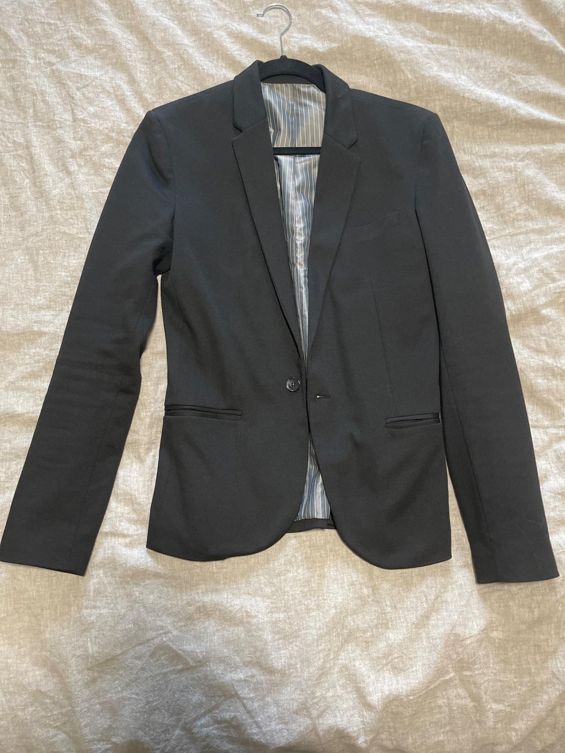 Black suit jacket hanging on a hanger over beige fabric. Inside with white and grey vertical striped lining.