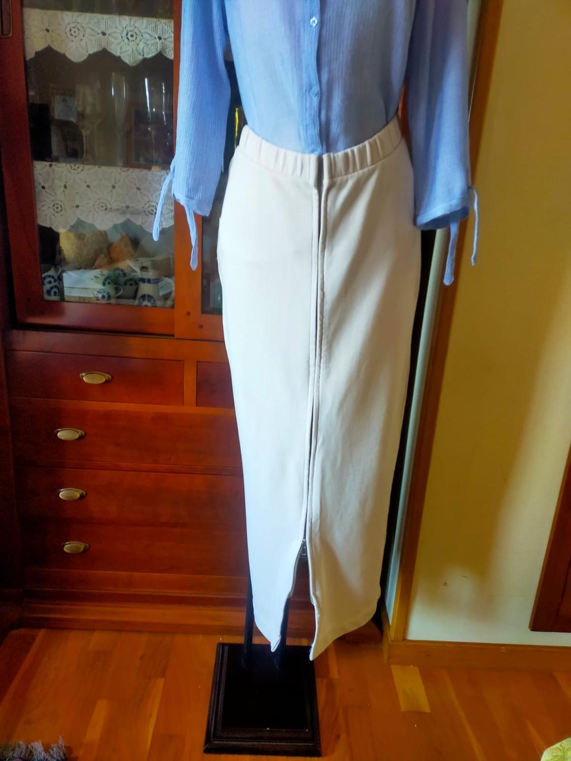 Mannequin with a long-sleeved blue blouse and a fitted white skirt with a front slit. Background of wooden furniture and parquet flooring.