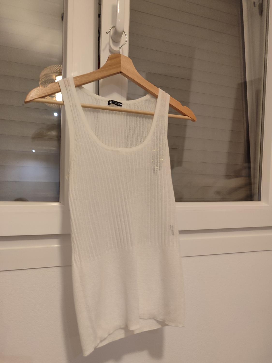White sleeveless top hanging on a wooden hanger in front of a window with closed blinds. Indoor setting.