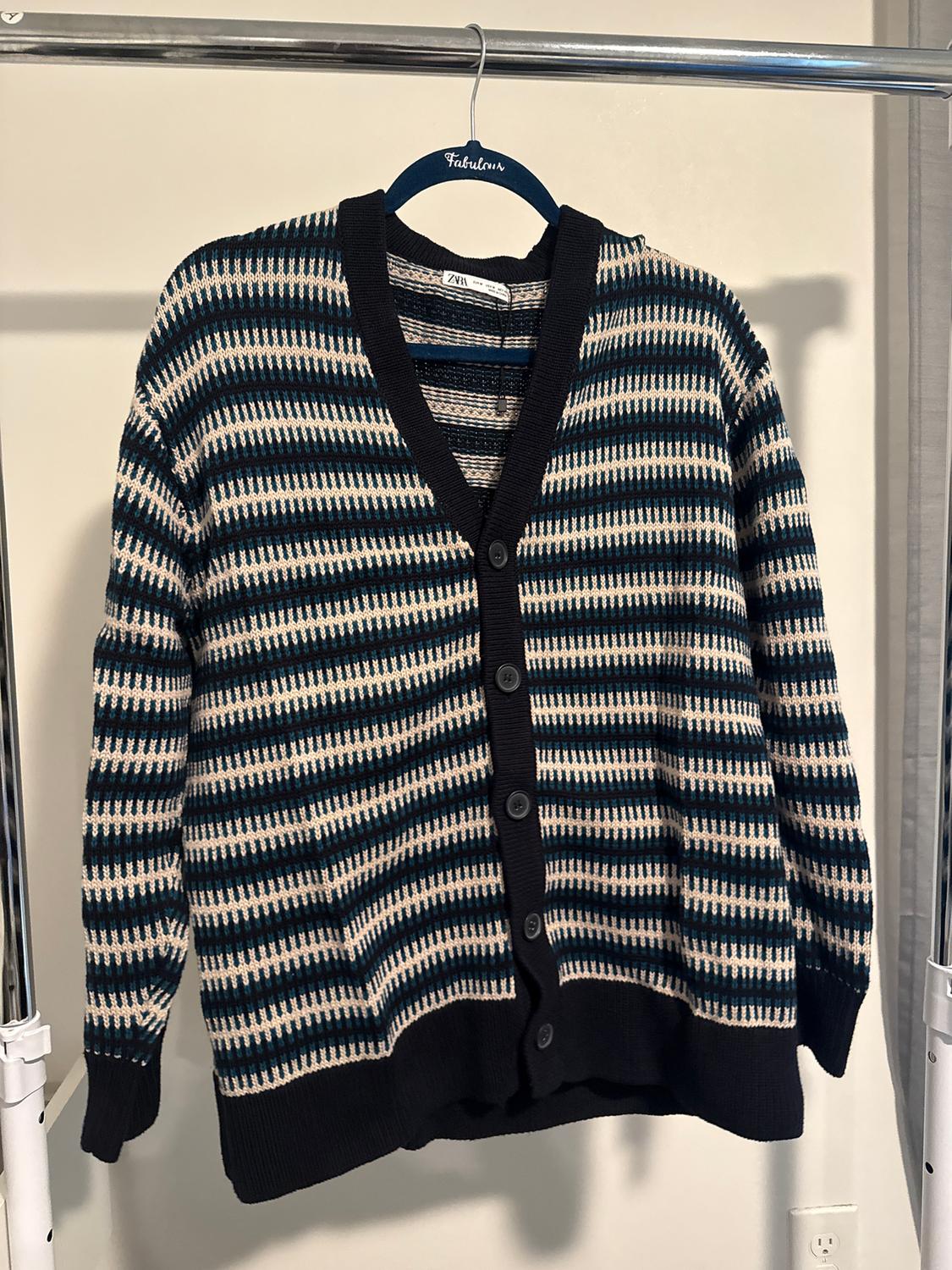 Knitted cardigan with a striped pattern in black, white, and blue, hanging on a blue hanger with the word "Fabulous".