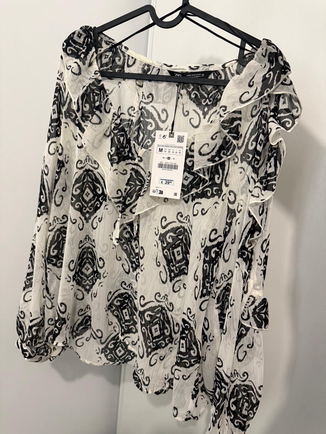 White blouse with black print hanging on a hanger. Visible tag with price of €29.95. White background.
