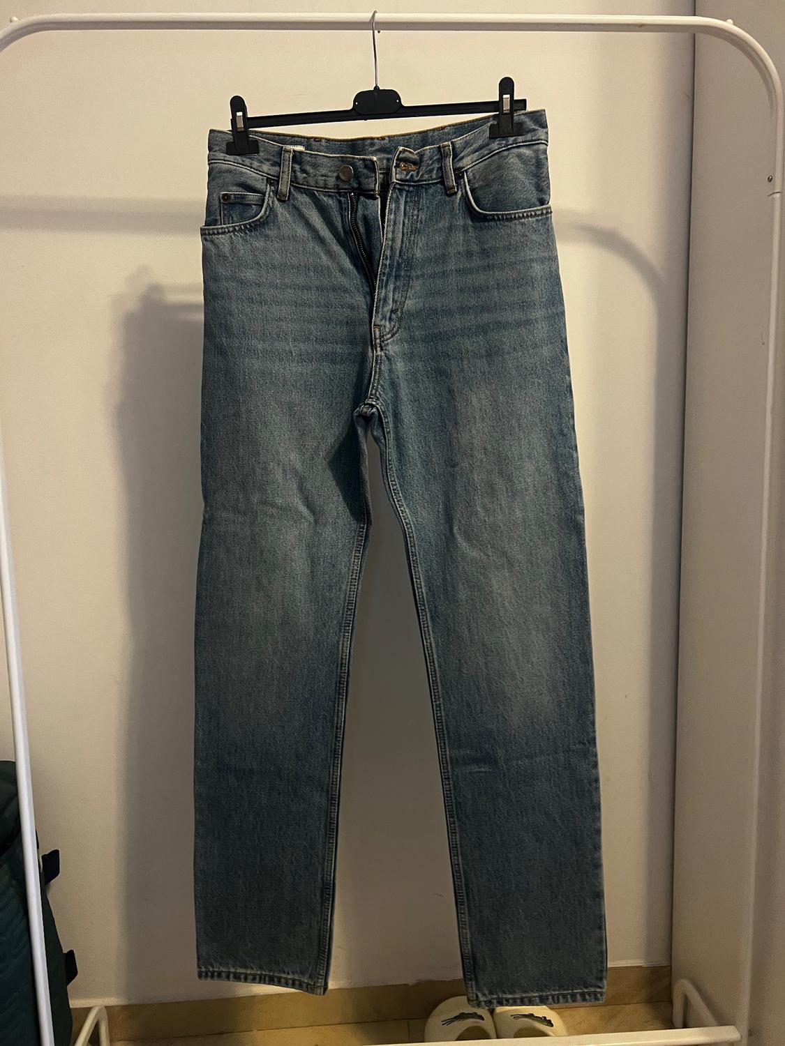 Blue jeans hanging on a black hanger against a white background. Bottom of a white shoe visible in the bottom corner.