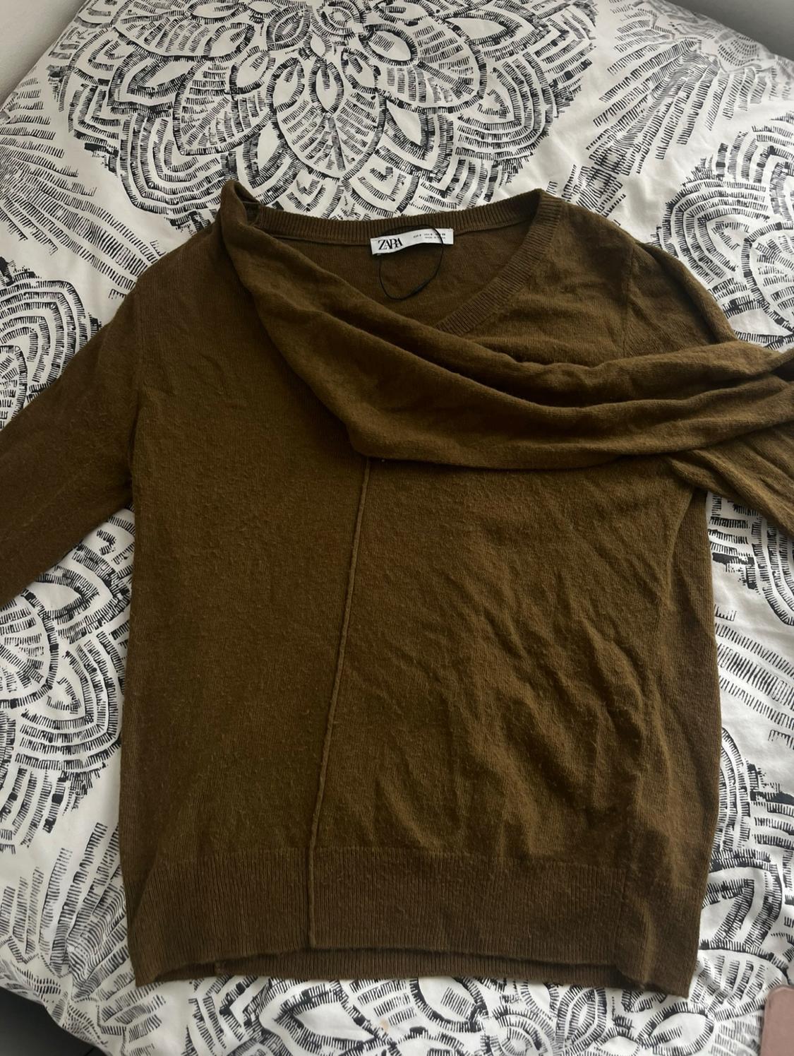 Brown knitted jumper with a draped neckline on a bed with a black-and-white patterned bedspread. ZARA text on the label.