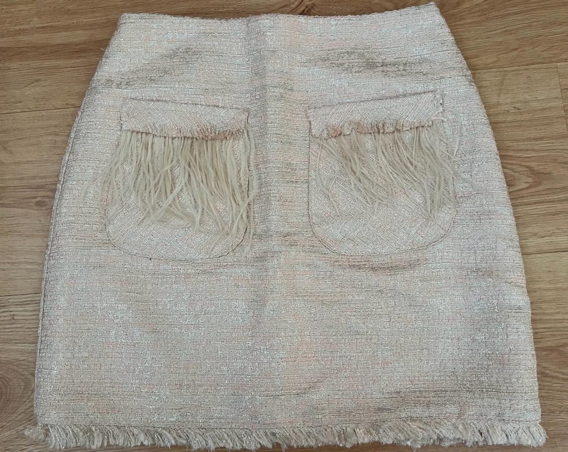 Light-toned, shimmery mini skirt on a wooden floor. Features two front pockets with feather detailing and a frayed hem.