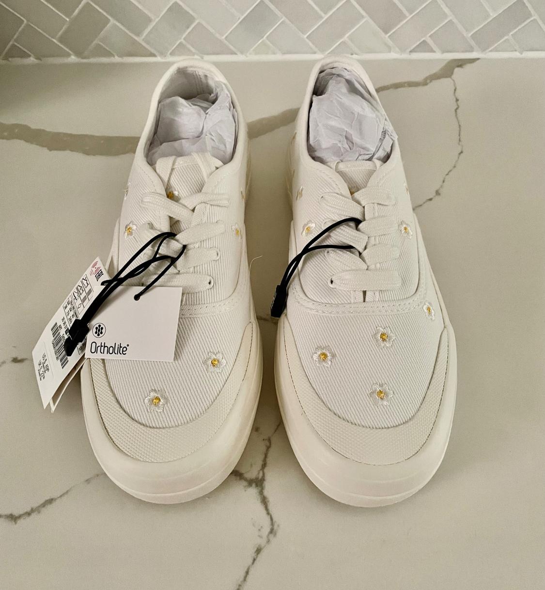 White trainers with embroidered floral details and swing tags on a marble surface. Visible text: "Ortholite".