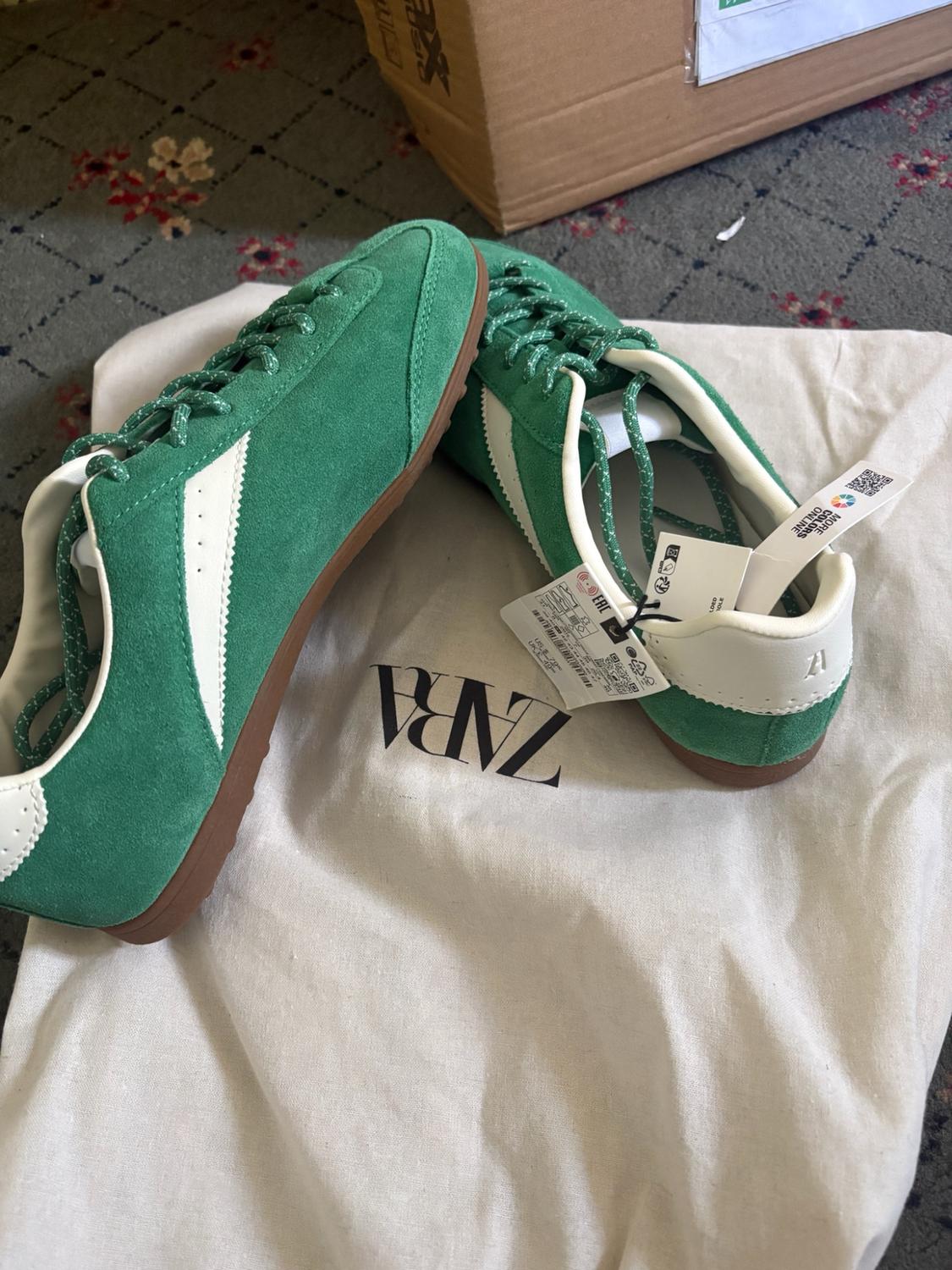 Green and white trainers on a fabric bag with the text ZARA. Label with the text MORE COLORS ONLINE. Casual setting with a cardboard box.