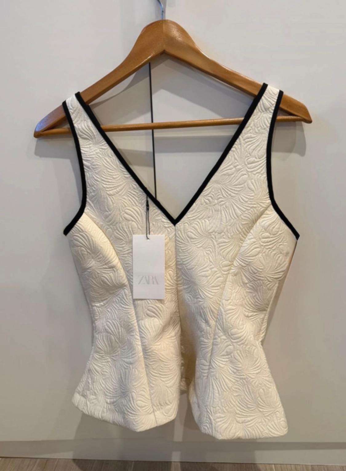 White top with a floral texture and black trim, hanging on a wooden hanger. Visible label with the text "ZARA".