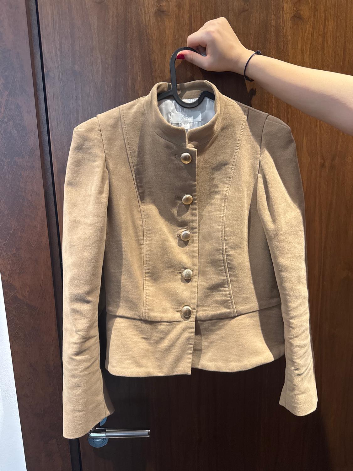 A beige high-necked jacket with large buttons, hanging on a hanger held by a hand over a wooden door.