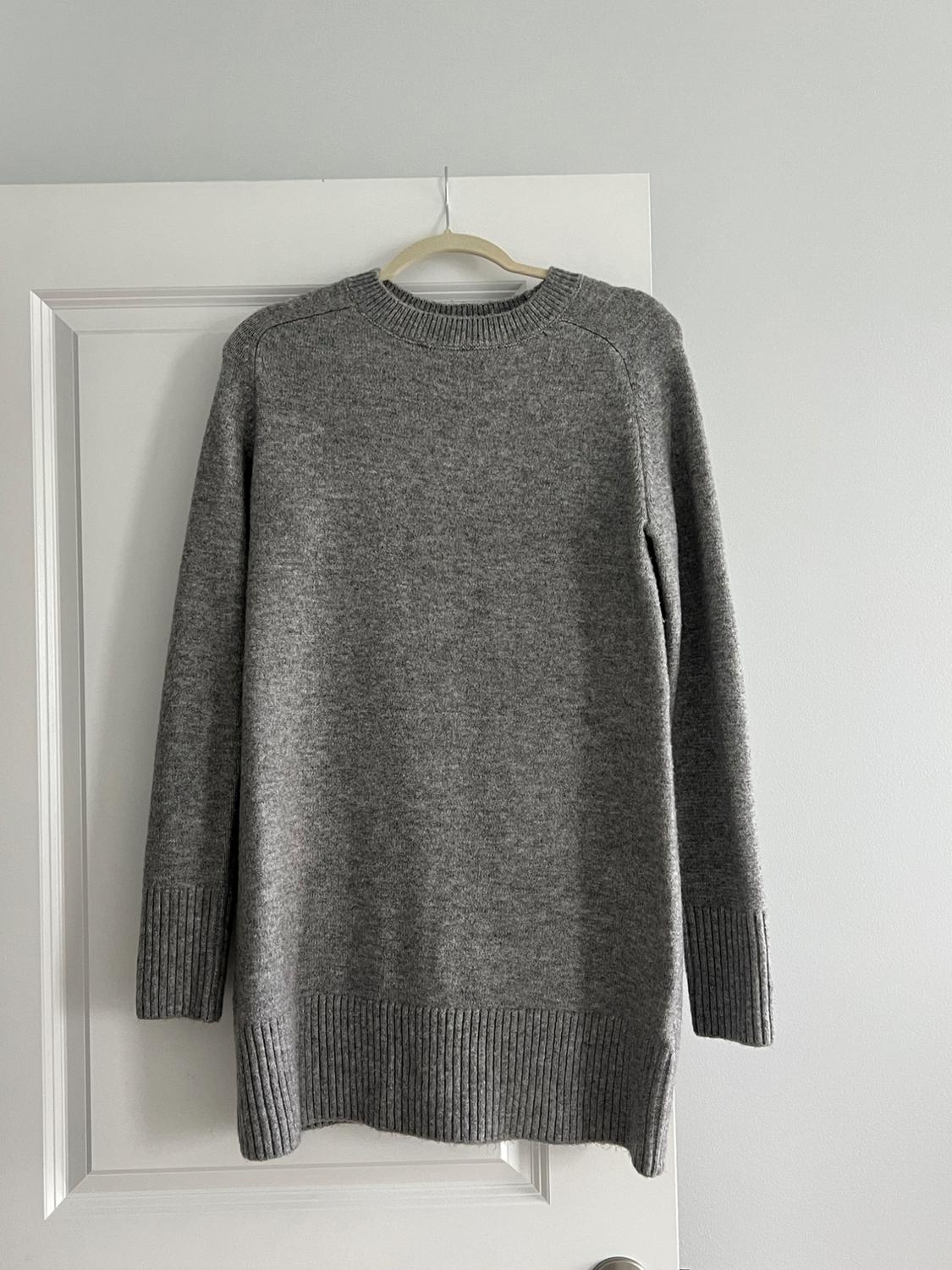 Gray knitted sweater hanging on a white door. Simple design with a crew neck and long sleeves. Minimalist setting.