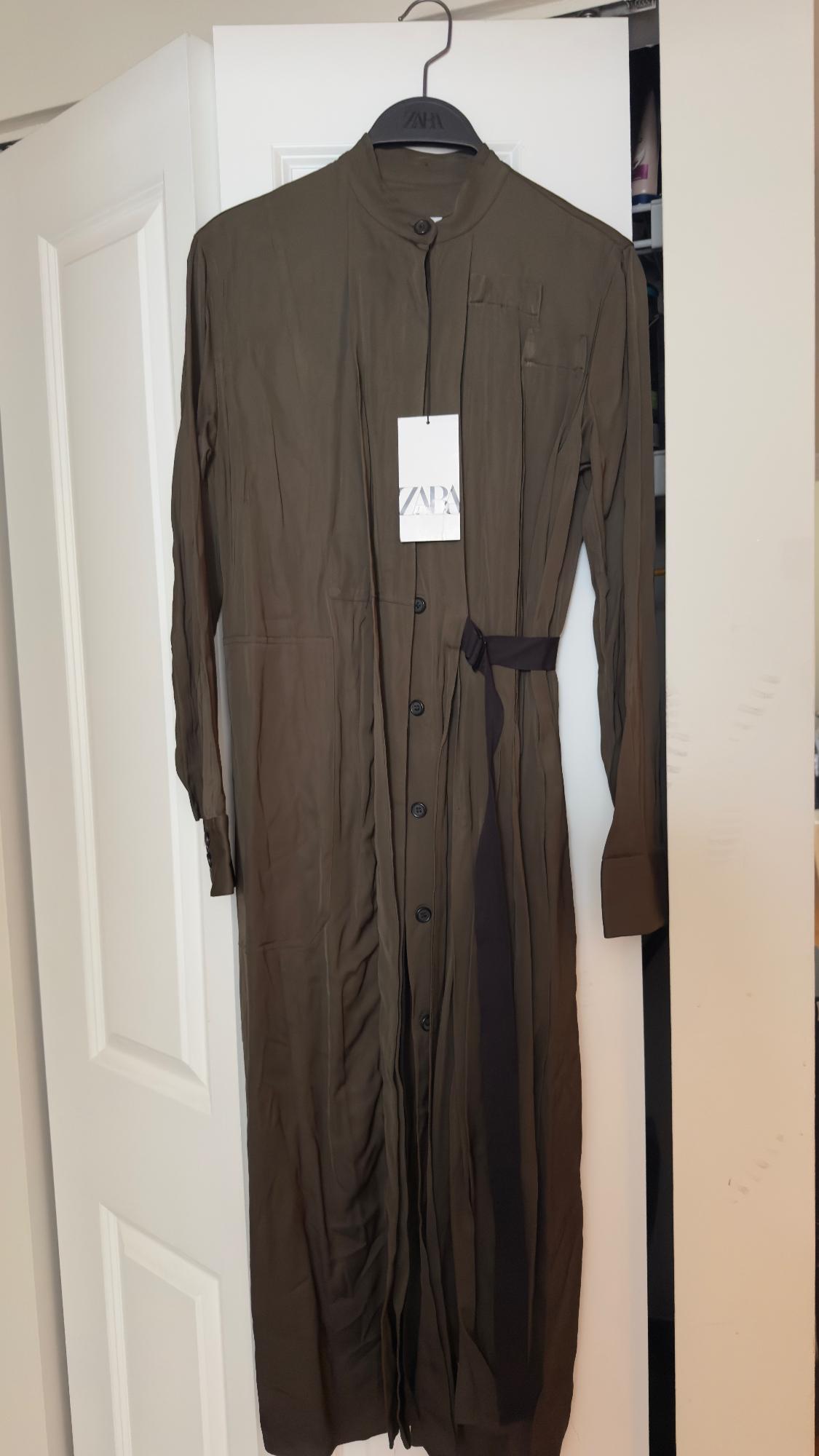 Long brown dress with buttons and a black belt, hanging on a black hanger. Visible tag with text "ZARA". White door in the background.