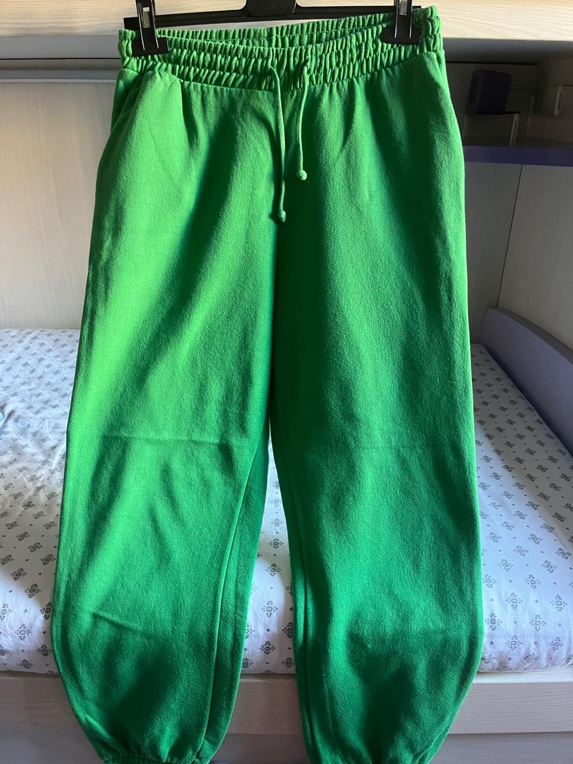 Green tracksuit bottoms with an elasticated waist and drawstring, hanging on a hanger over a bed with patterned sheets. Casual style.
