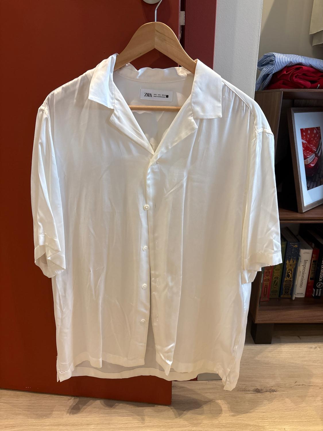 White short-sleeved shirt hanging on a wooden hanger, against a red background. In the background, a bookshelf with books and folded clothes.