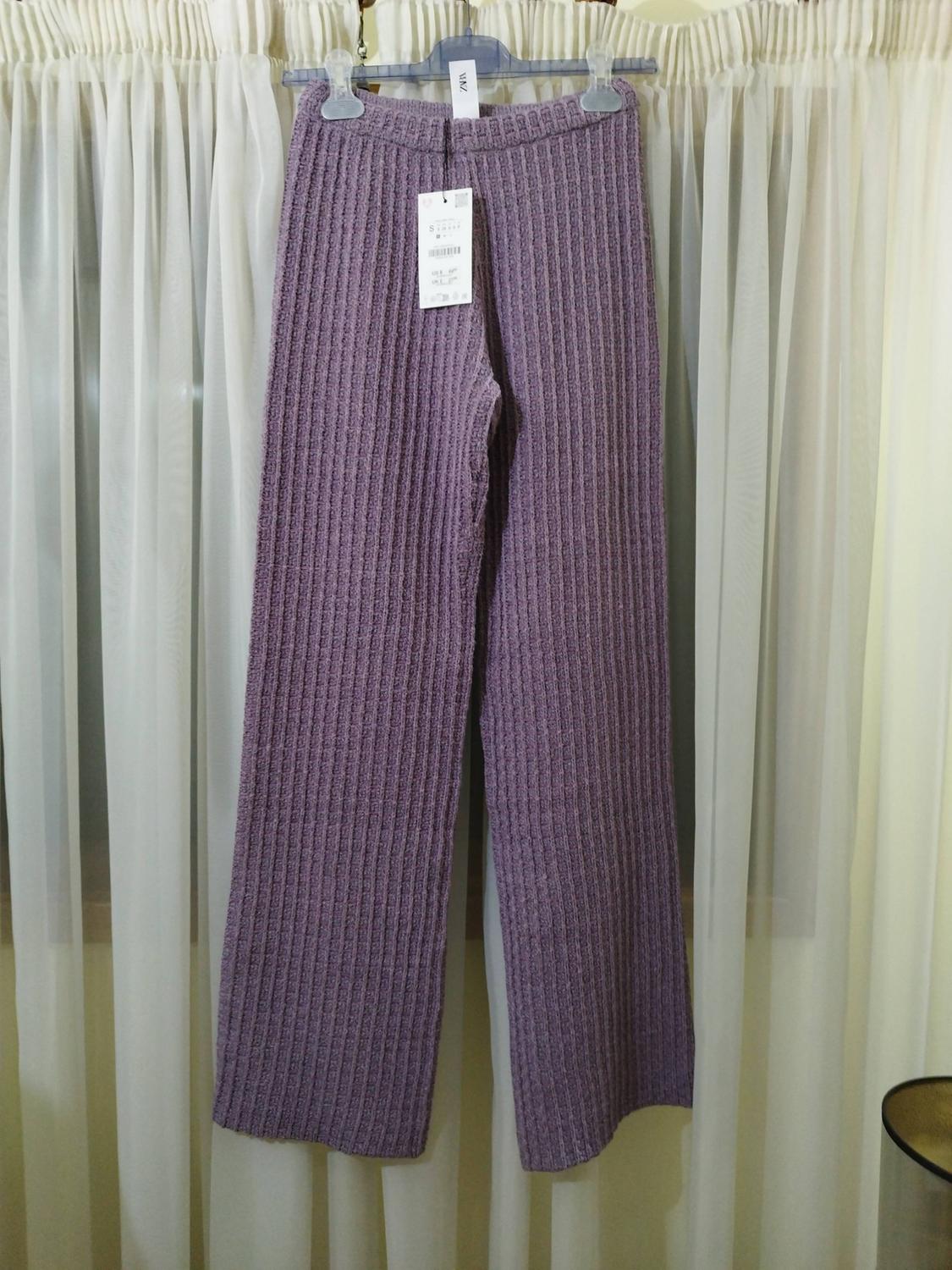 Purple ribbed-knit trousers hanging on a hanger with the text ZARA and a tag, in front of sheer white curtains.