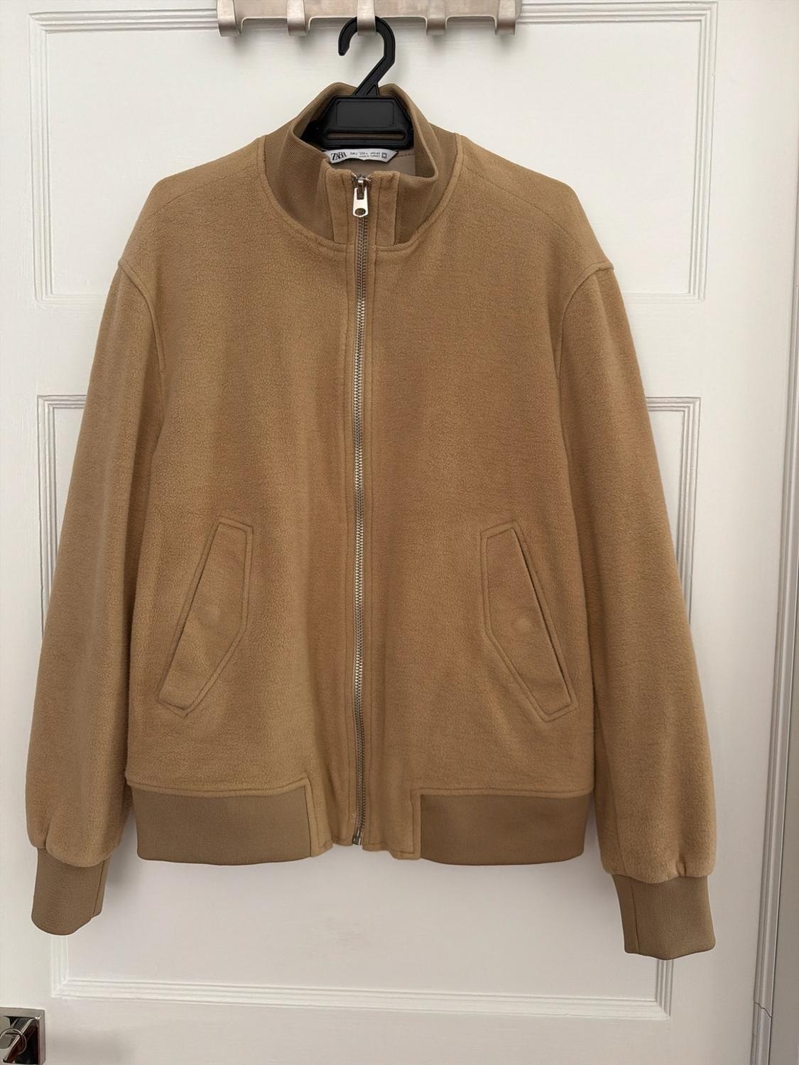 Camel jacket with zipper, high neck, and pockets, hanging on a black hanger over a white door. Text: ZARA.
