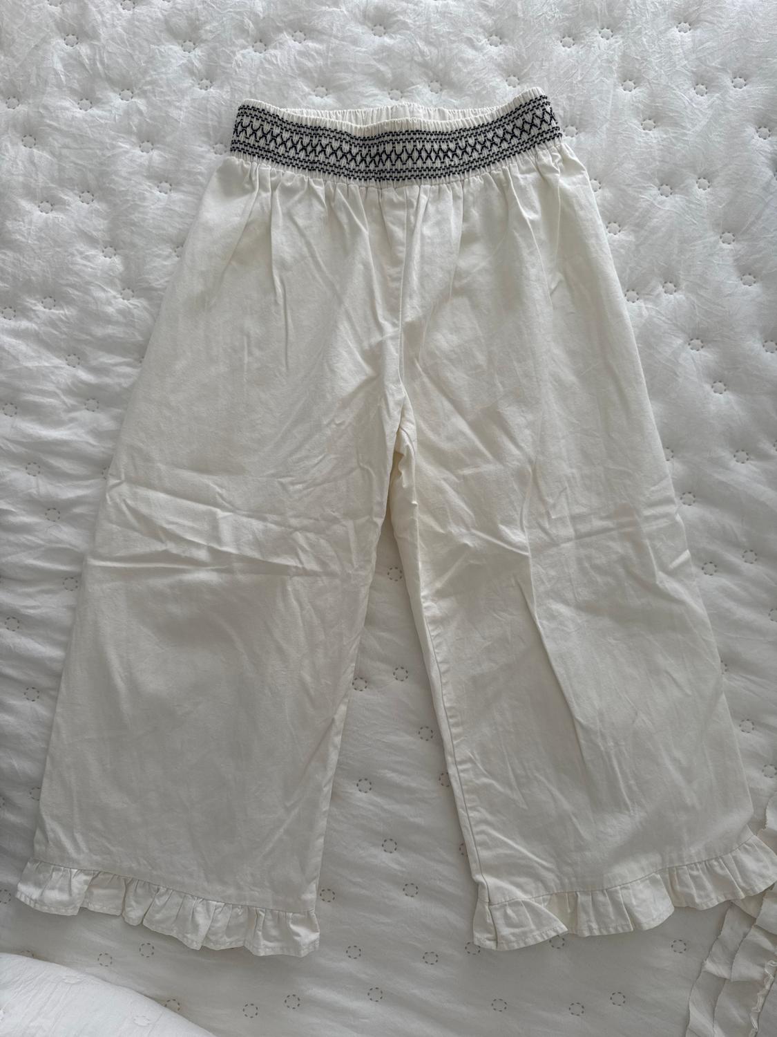 White cotton trousers with a black elasticated waist and ruffled detailing on the hems, on a white quilted bedspread.