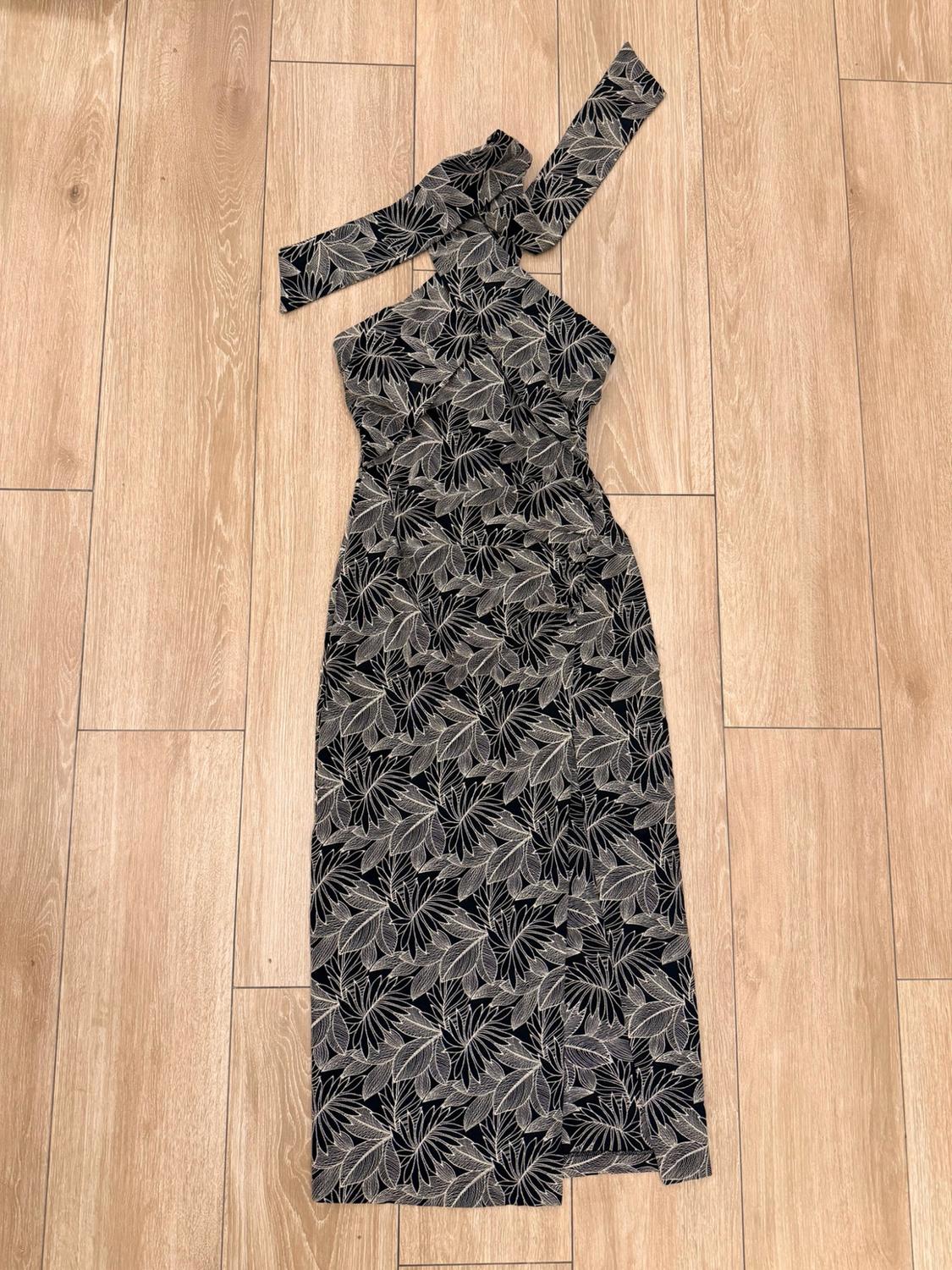 Black dress with a white leaf print, a halter neck with a tie and a slit in the skirt, laid out on a wooden floor.