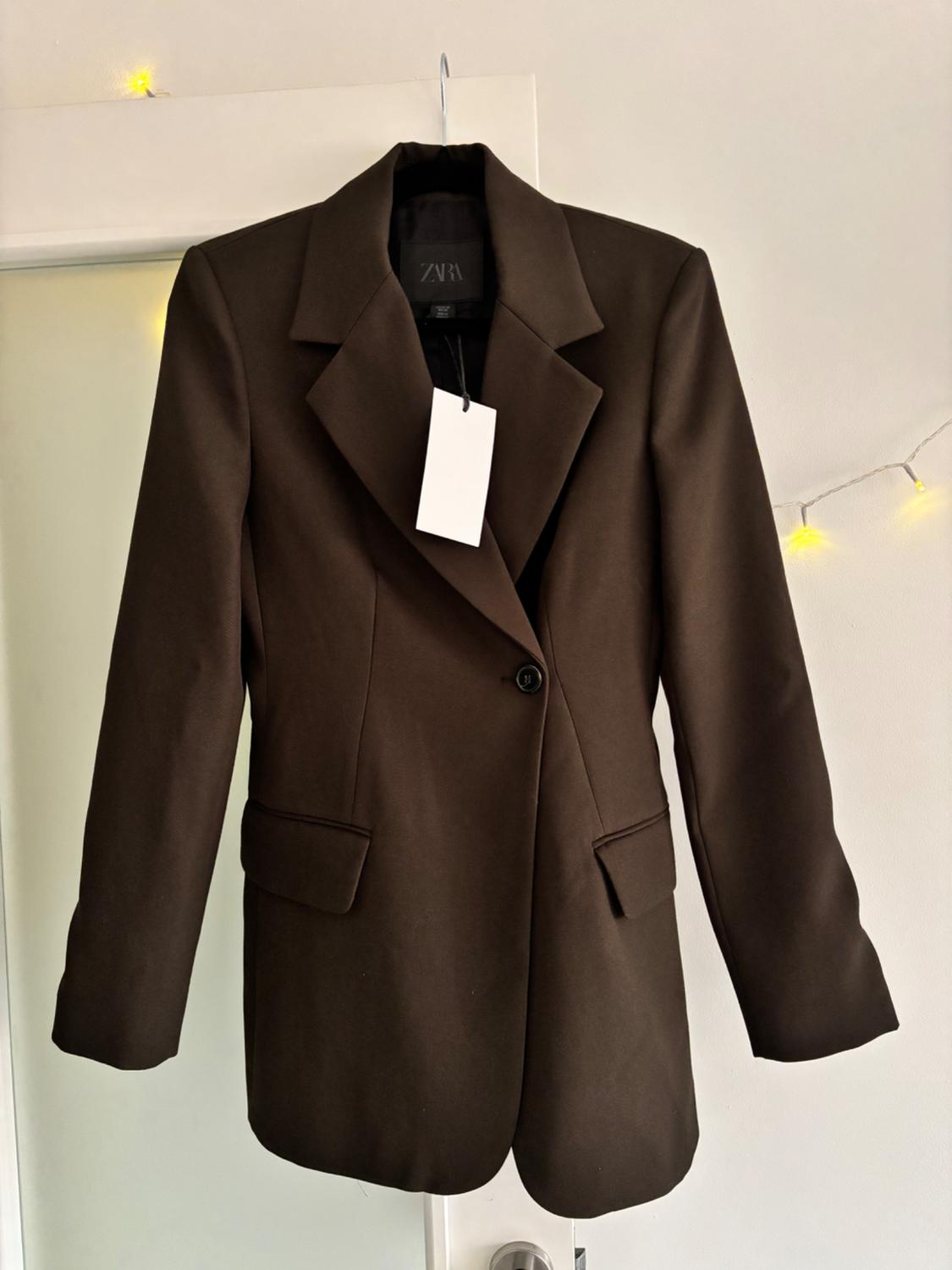 Brown, single-breasted jacket hanging on a hanger, with a visible white tag. Background with decorative yellow lights.