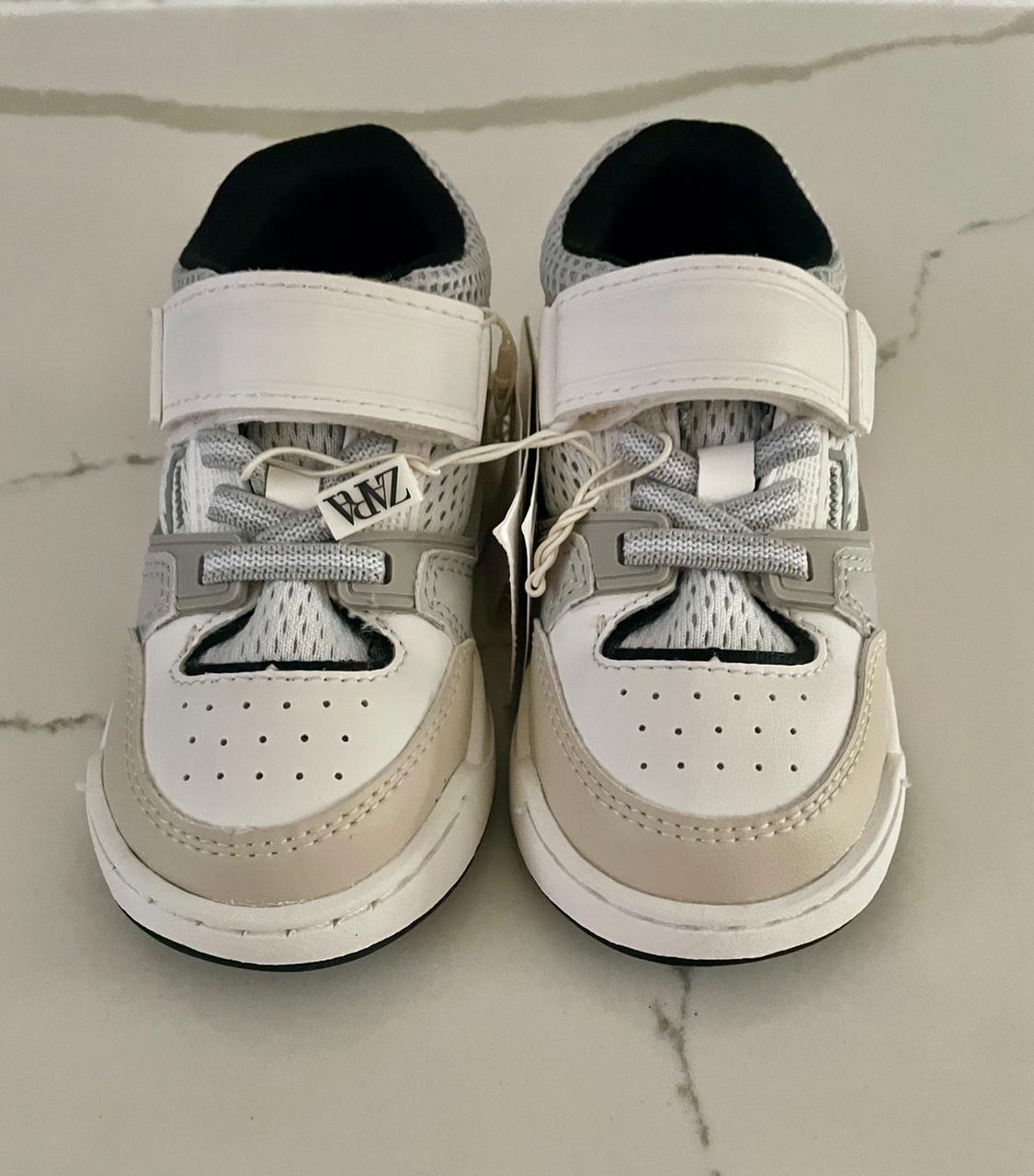 Sports footwear in white and grey tones with an adhesive fastening and "ZARA" label, on a light marble surface.