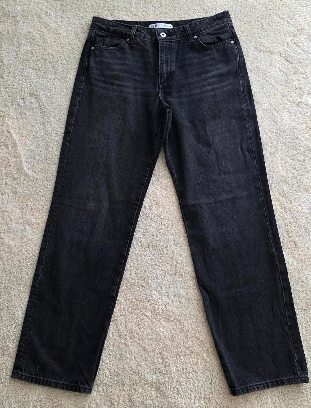 Black straight-cut jeans on a beige rug. Visible label on the waistband with the text "ZARA".