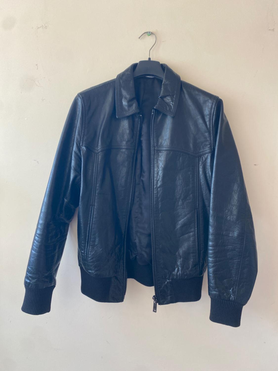 Black leather jacket hanging on a hanger against a beige wall. It has a front zip and ribbed cuffs.