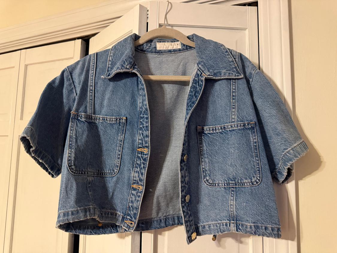 Light blue denim jacket with short sleeves and two front pockets, hanging on a hanger in front of a white closet.