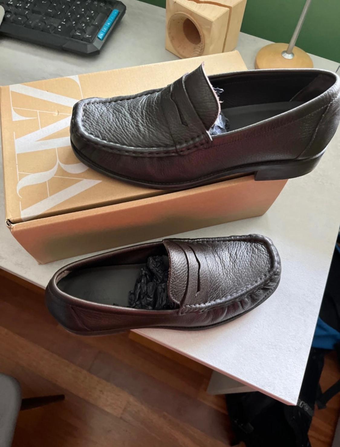 Pair of dark brown leather loafers. One is on a box bearing the text ZARA and the other is on a desk.