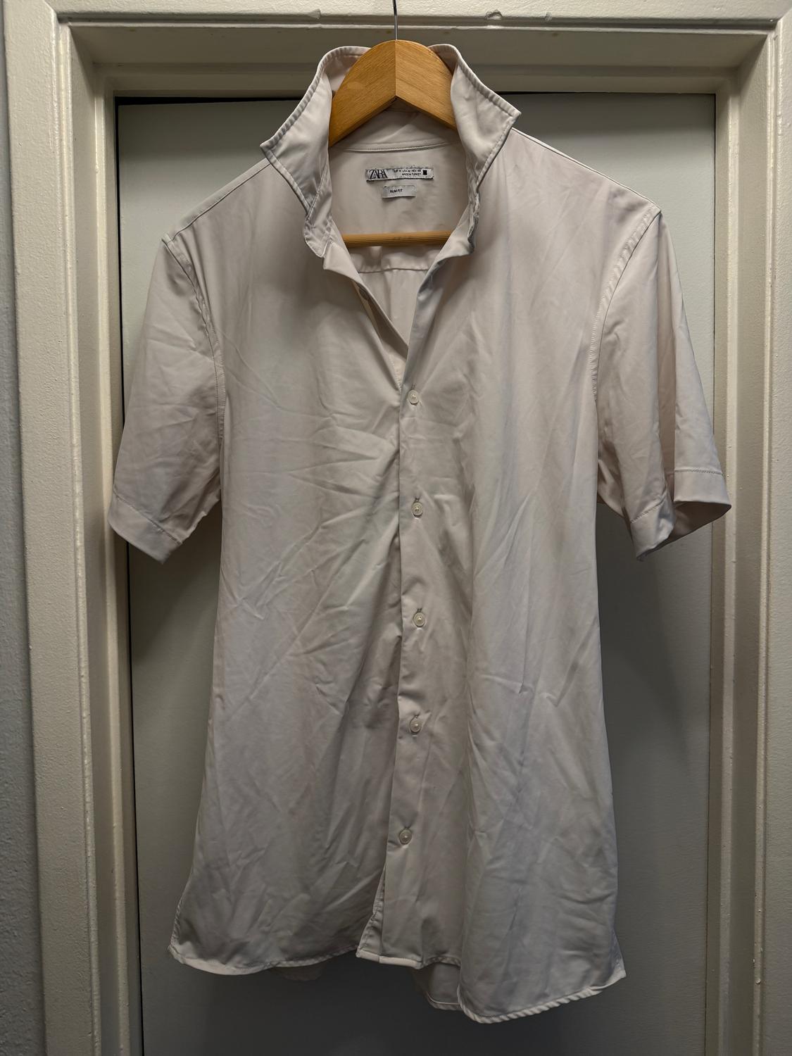 Short-sleeved white shirt hanging on a wooden hanger. The collar is up and the fabric has visible wrinkles.