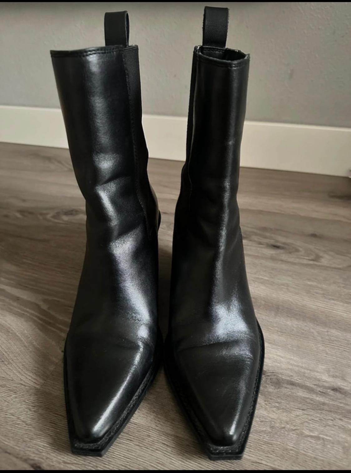 Black pointed ankle boots with pull tabs, on a wooden floor in front of a grey wall, with a casual and relaxed style.