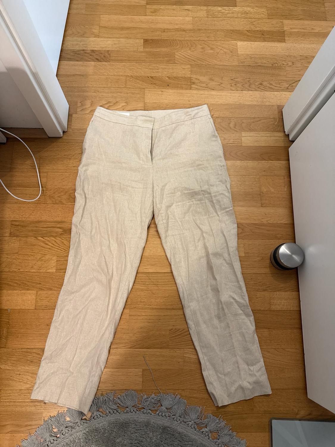 Light beige trousers laid out on a wooden floor. White door frames and a grey rug stand out in the composition.