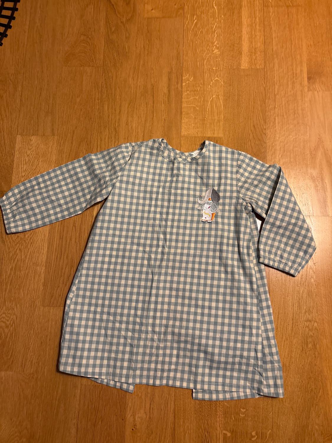 Child's blue and white checked dress on a wooden floor. It has a small mouse embroidery on the chest.