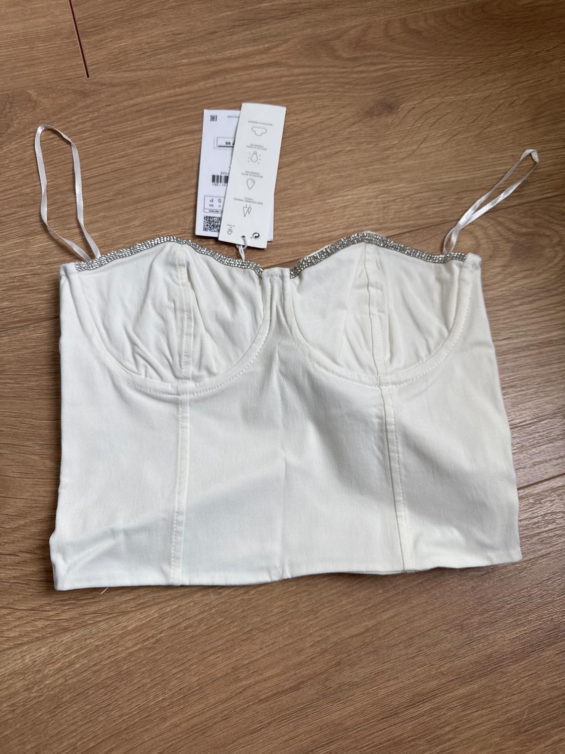 White top with thin straps and sparkly details on the top edge, on a wooden floor. Visible label with text.