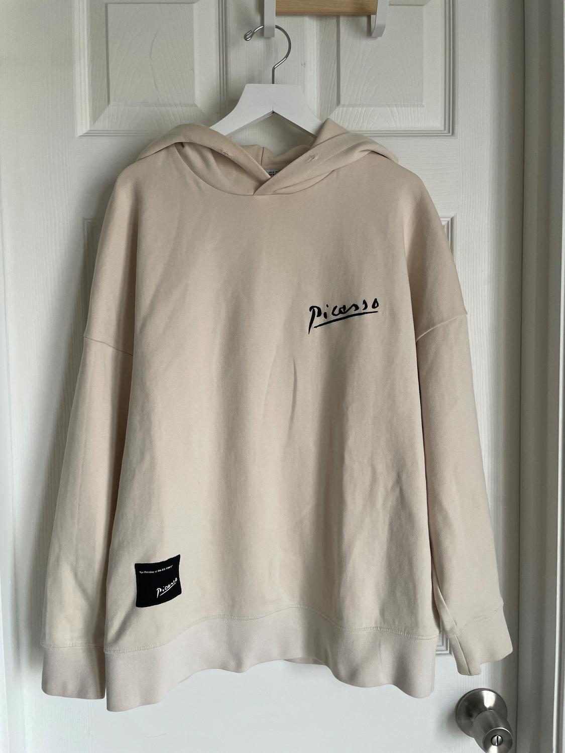 Beige hoodie hanging on a white door. Text "Picasso" on the chest and a black tag on the hem.