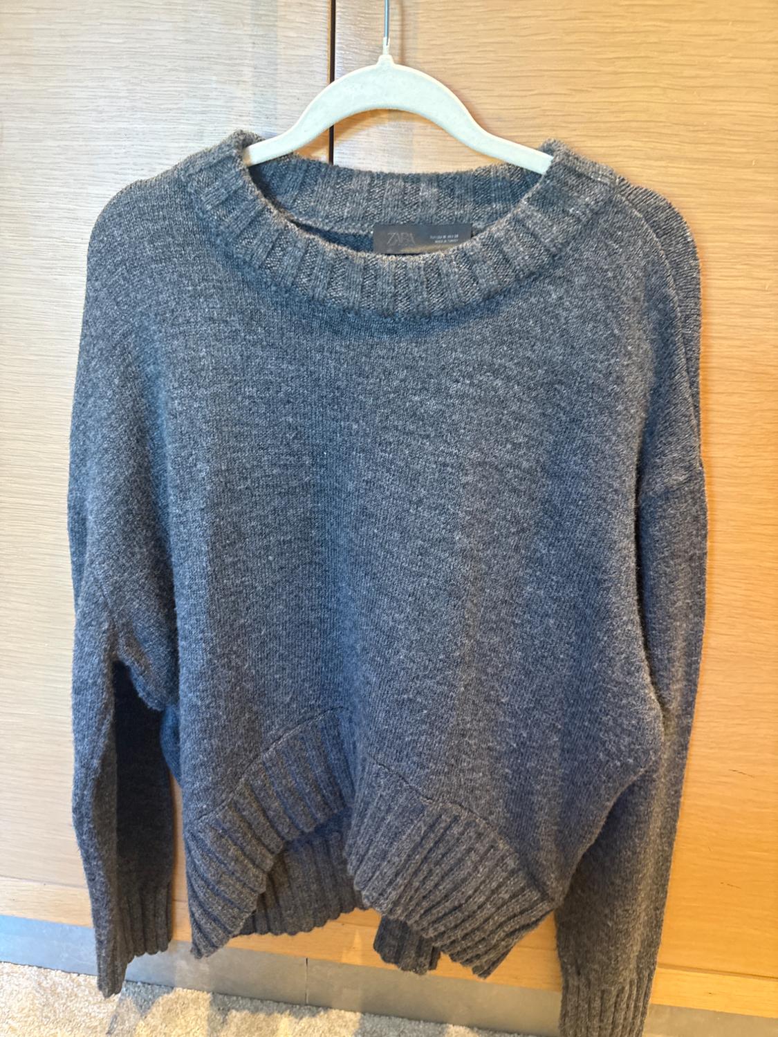 Chunky knit grey jumper hanging on a white hanger against a light wooden background. Label visible on the collar.