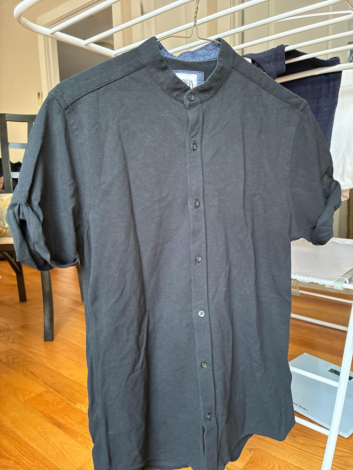 Black shirt with a ZARA label, rolled-up short sleeves, and a mandarin collar, hanging on a white clothesline over a wooden floor.