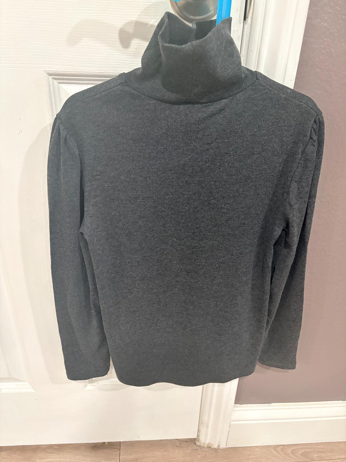 Dark gray turtleneck sweater hanging on a hanger, over a white door. Long sleeve and plain knit.