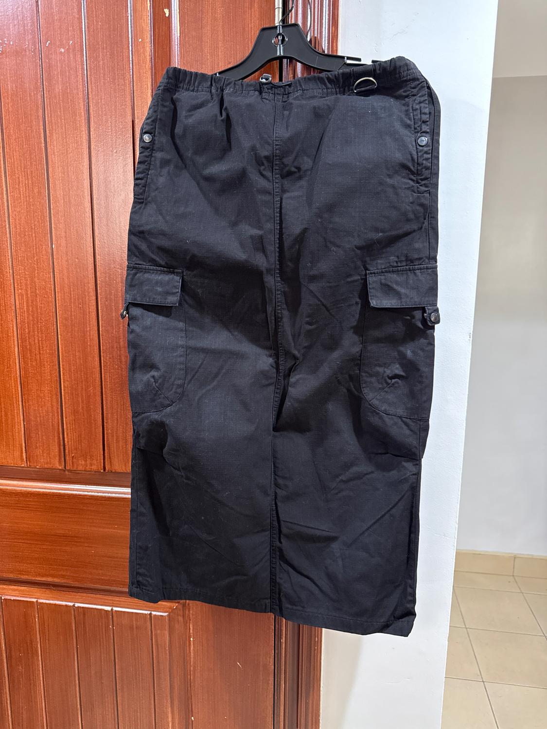 Black skirt hanging on a hanger in front of a wooden door. It has side pockets with flaps and buttons.