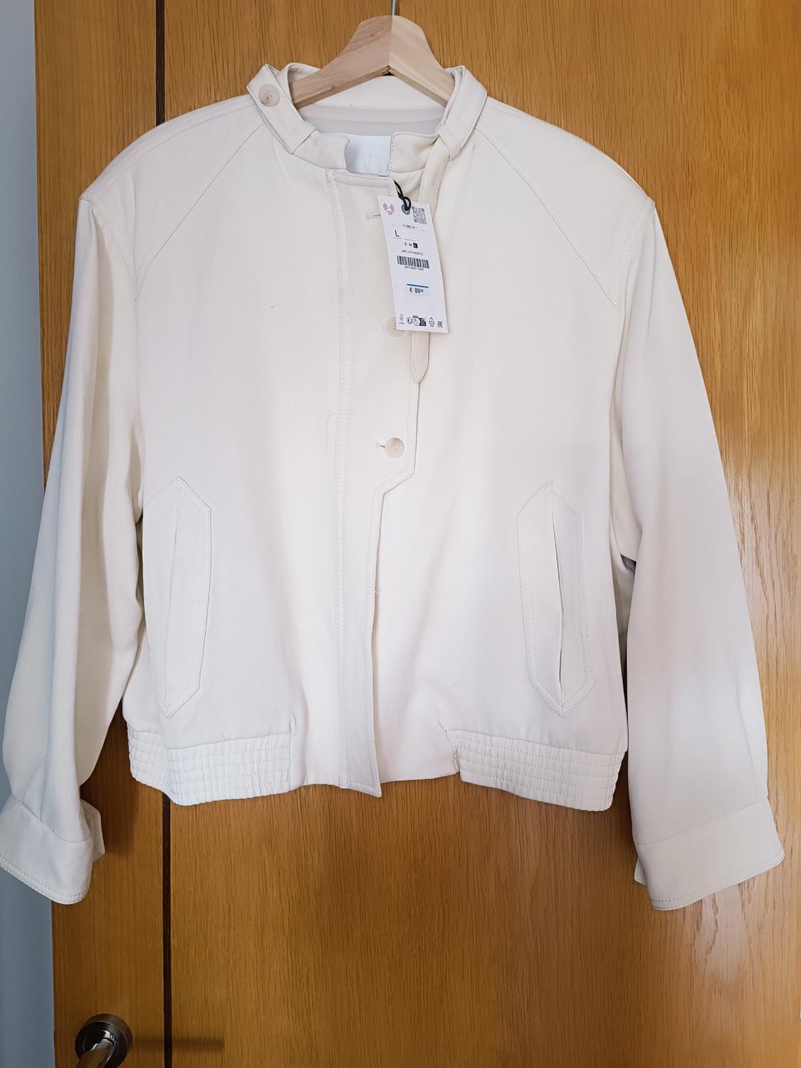 White jacket hanging on a wooden hanger over a wooden door. Visible label with barcode and size L.