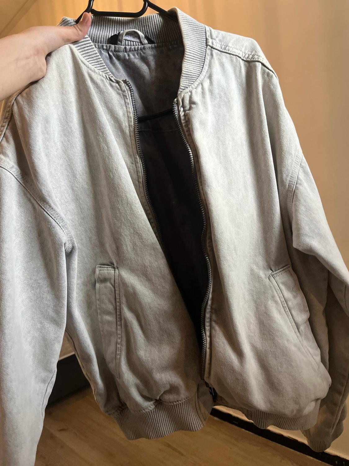 Hand holding a light grey bomber jacket on a black hanger. Open design with zip fastening, side pockets and ribbed trims.