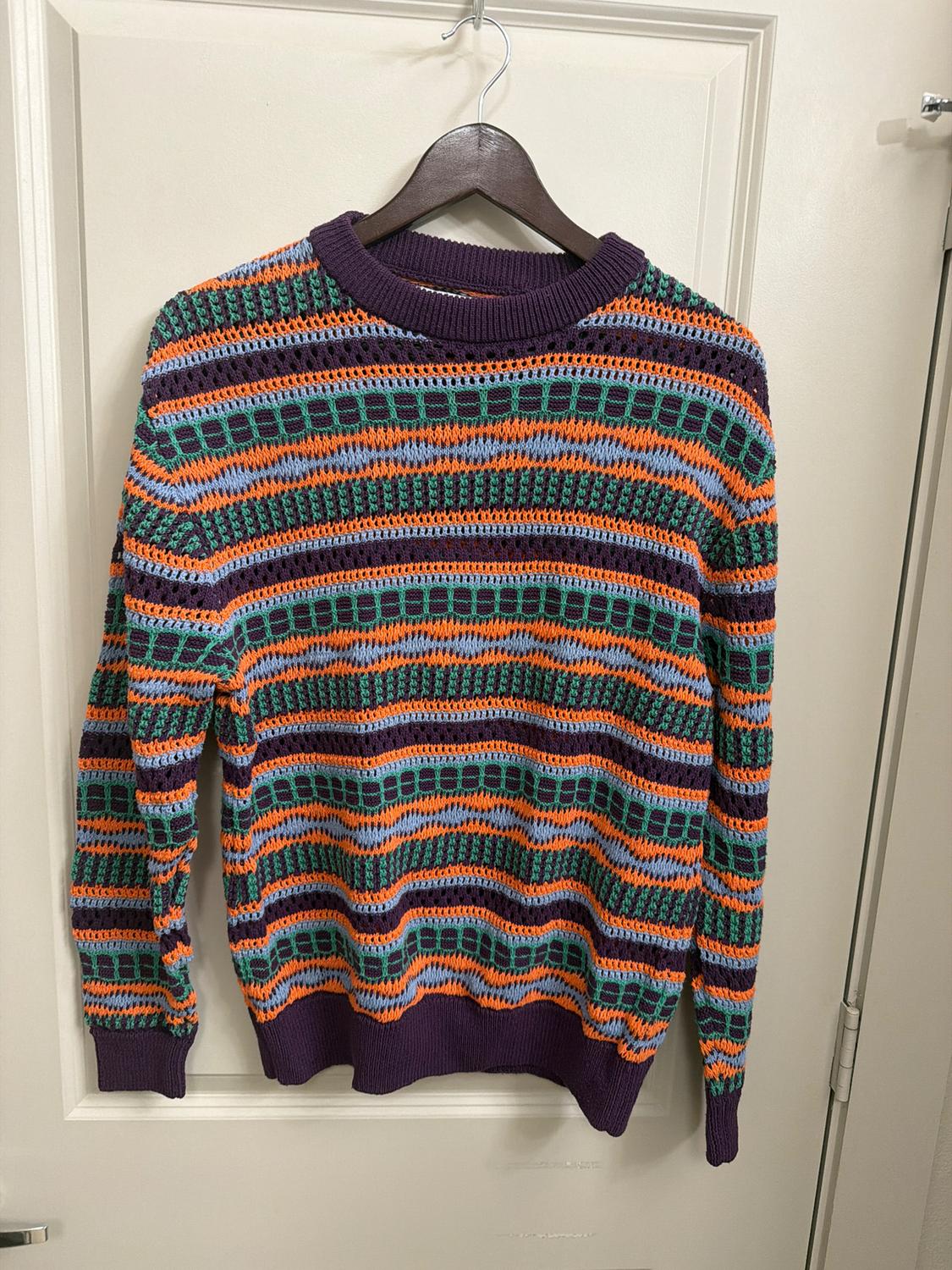 Knitted jumper hanging on a hanger, with a multi-coloured striped pattern in shades of purple, orange, green, and blue. White door background.
