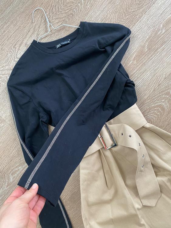 Black long-sleeved shirt with visible stitching and beige pants with a metallic buckle belt on a wooden floor.