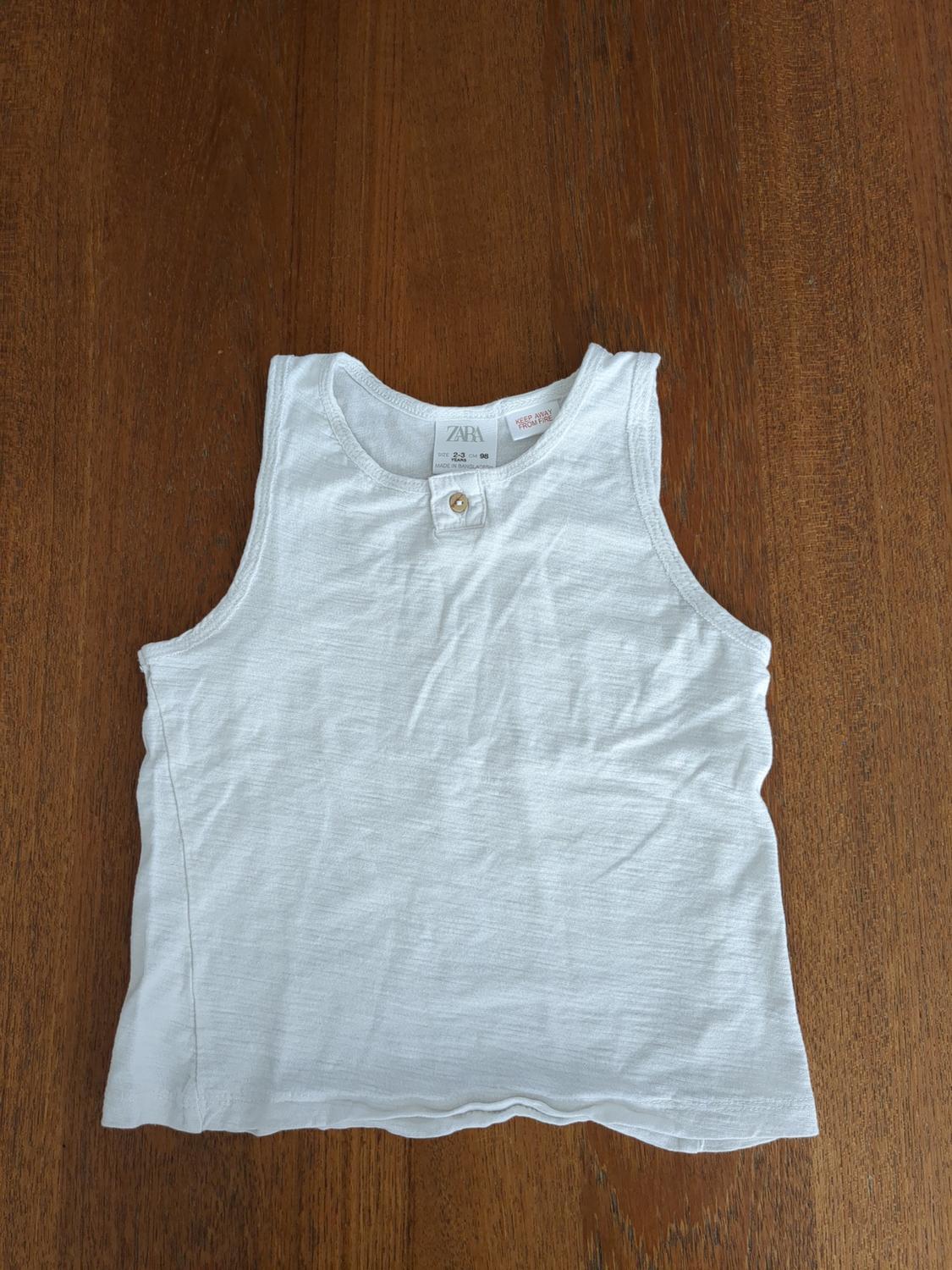 Boy's white sleeveless t-shirt on wooden table. Visible tag with text: "ZARA, 2-3 years, 98 cm, Made in India".