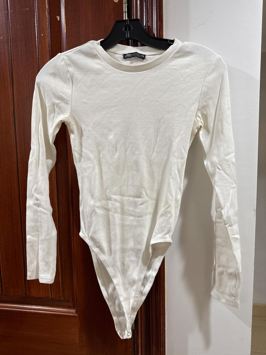 Long-sleeved white bodysuit hanging on a black hanger on a wooden door.