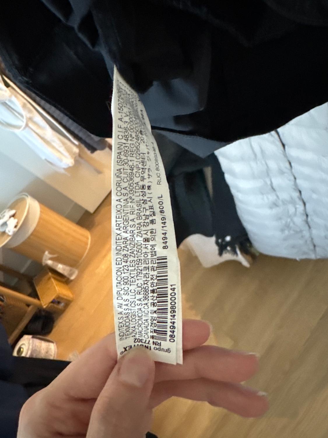Garment tag with multi-language text held by a hand. Background with wooden furniture and hanging clothes.