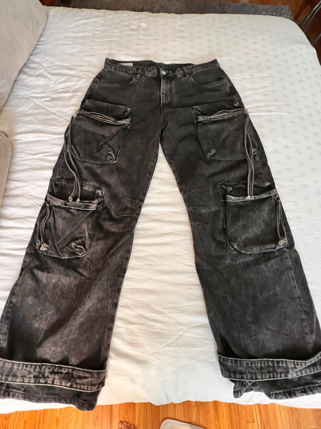 Washed dark gray cargo jeans with multiple pockets, zippers, and long straps, laid out on a white bed.