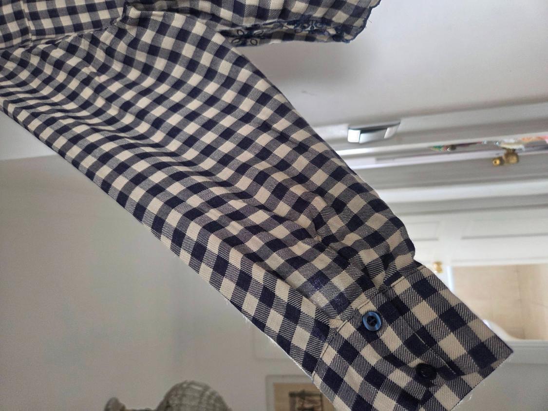 Detail of a navy and white checked shirt sleeve. The cuff with a blue button is visible in a relaxed, out-of-focus indoor setting.