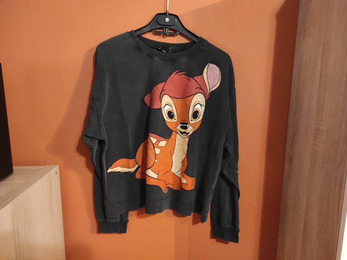Black sweatshirt with a cartoon deer graphic on the front, hanging on a hanger against an orange wall.