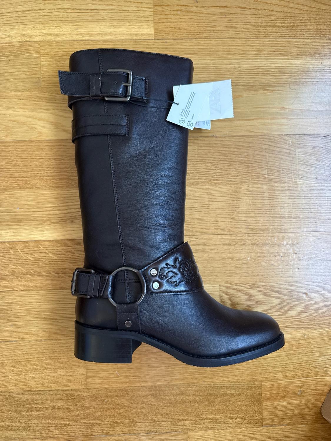 Dark brown leather high boot with buckles, embossed detail at the ankle and ZARA label, resting on a wooden floor.