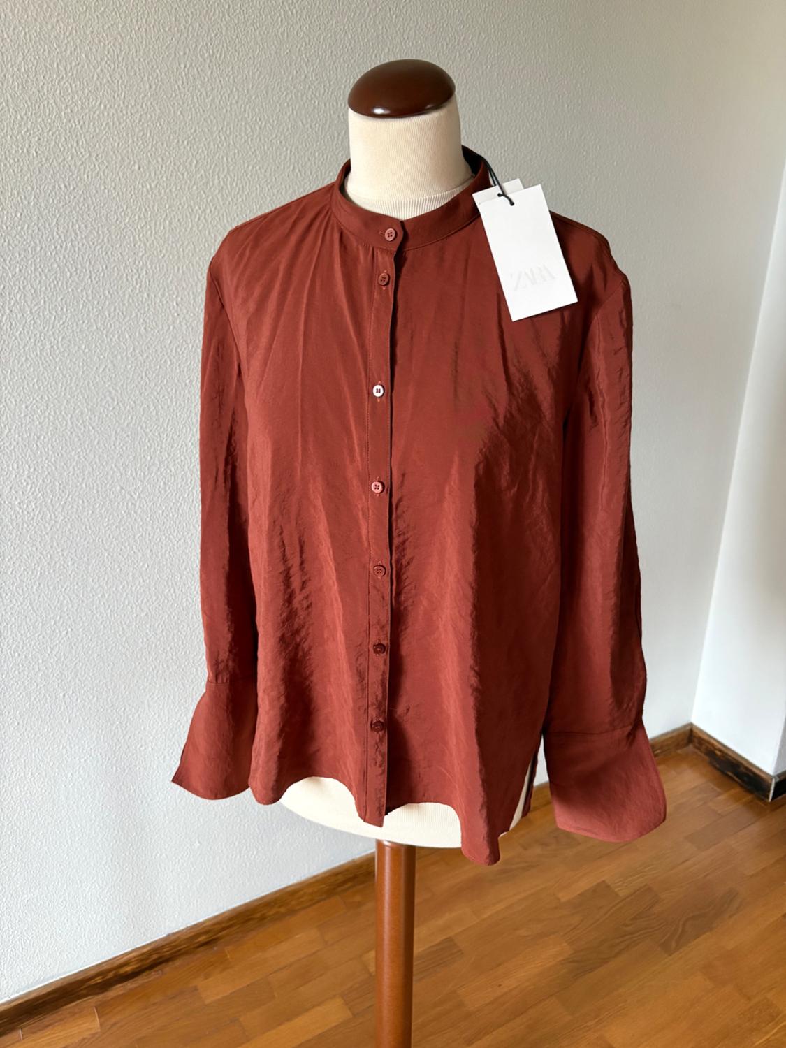 Terracotta-coloured long-sleeved shirt with a mandarin collar on a mannequin. Label with ZARA text. Interior background with a white wall and wooden floor.