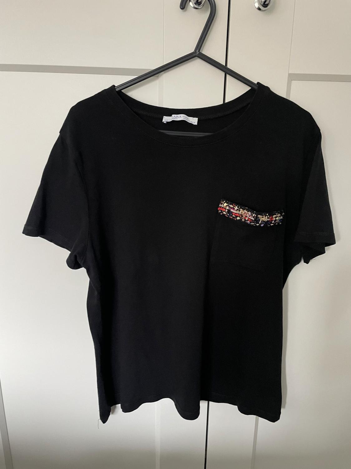 Black t-shirt hanging on a hanger, with a pocket decorated with colourful fabric. White doors in the background. Label visible.