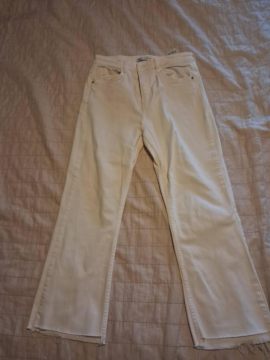 White denim jeans with a frayed hem on a bed with a beige bedspread. Inside label bearing the text ZARA.